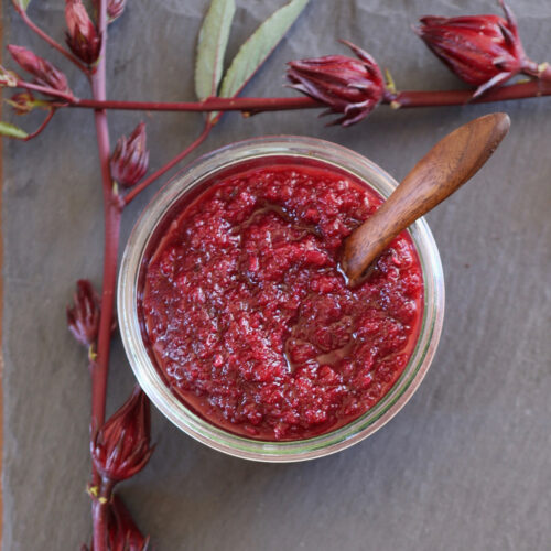 Hibiscus Chutney in a glass jar surrounded by fresh hibiscus calyces.